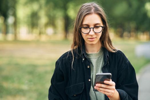 Young woman with glasses reading a message on her phone while standing in the park. High quality photo