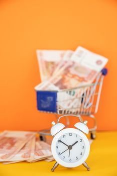 Mini shopping cart with 100 dollar banknotes inside and mini alarm clock on yellow background. Trolley and money.