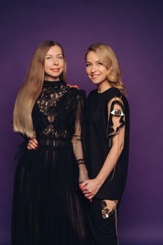 Two sisters pose in the studio on a purple background. 2 beautiful women in black fancy dresses gathered for a party