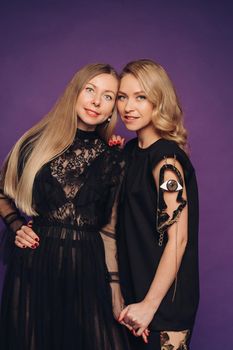 Two sisters pose in the studio on a purple background. 2 beautiful women in black fancy dresses gathered for a party