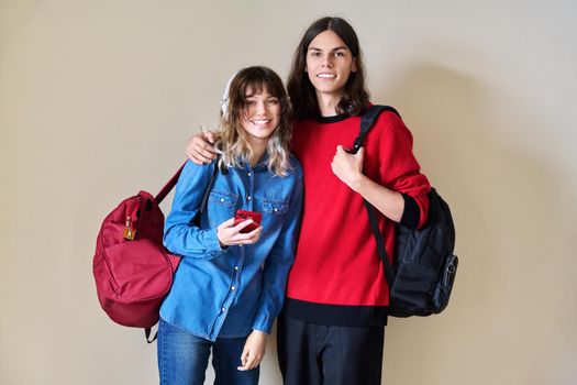 Portrait of teenage guy and girl together, smiling positive looking at camera. Friends students with backpacks on background of light wall. Youth, college, high school, education, young people concept