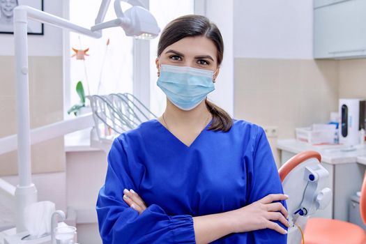 Portrait of confident female dentist doctor in office looking at camera. Young female in medical protective mask in blue uniform. Dentistry, medicine, health care, profession, stomatology