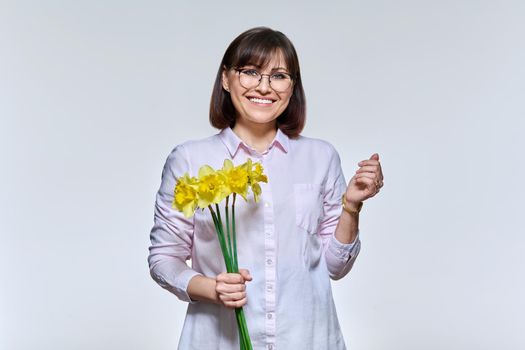 Portrait of beautiful middle aged woman with bouquet of flowers looking at camera on light studio background. Charming smiling mature woman. Beauty, holiday, joy, happiness, people concept