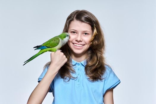 Smiling child girl with pet green quaker parrot looking at camera on light studio background. Animals, bird owner, childhood, child and pet friendship concept