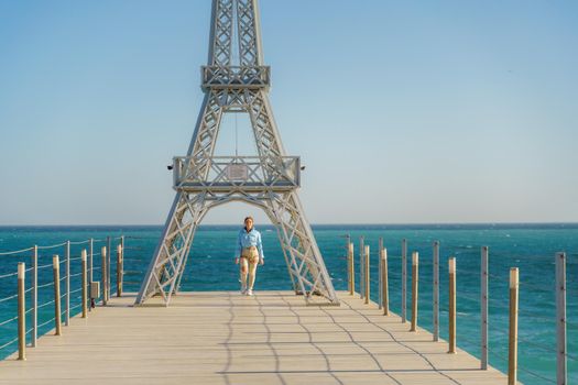 Large model of the Eiffel Tower on the beach. A woman walks along the pier towards the tower, wearing a blue jacket and white jeans