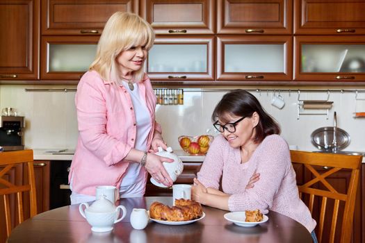 Two smiling happy mature women girlfriends in the kitchen together, having fun drinking tea chatting. Friendship, leisure, communication, 40s 50s age people concept