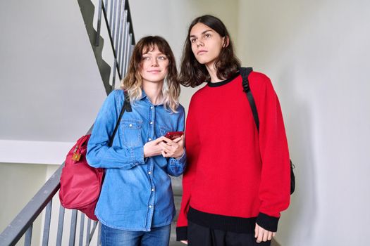 Portrait of teenage couple students 17, 18 years old with backpacks standing together on stairs inside looking away. Youth, lifestyle, friendship, young people concept