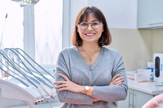 Portrait of smiling mature woman in dental clinic looking at camera. Confident female with crossed arms, dental office interior with equipment. Treatment, therapy, dental care, prosthetics, dentistry