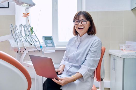 Portrait of dentist doctor sitting in office with laptop in her hands looking at camera. Mature smiling female nurse in dental clinic. Dentistry, medicine, health care, profession, stomatology concept