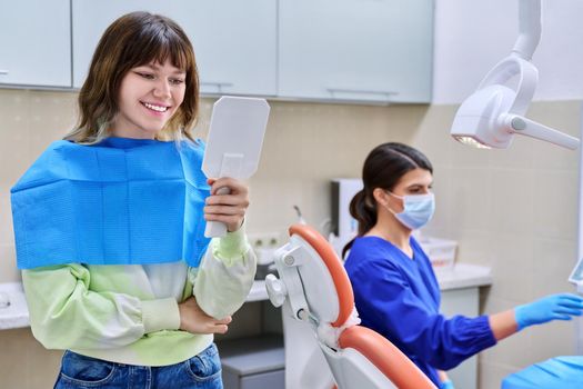 Teenage female looking at healthy teeth in mirror, in dental office, visit to dentist orthodontist. Treatment and care of teeth, teenagers, orthodontics, dentistry concept