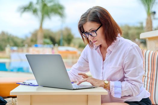 Beautiful business woman in her 40s working on a laptop remotely, sitting at an outdoor table in a resort hotel. Freelance work, business, middle aged people concept