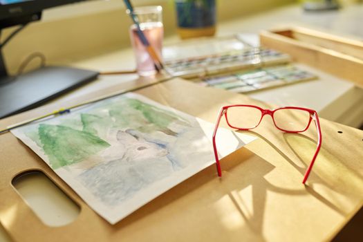 Drawing, artwork, hobby, education, creativity concept. Close-up of watercolor drawing, glasses, watercolor paints in box, brushes on the table, nobody