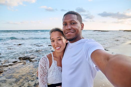 Happy couple in love taking selfie together on smartphone on beach, looking at camera. Vacation tourist trip to seaside resort, summer vacation, multicultural family, happiness, date, love concept