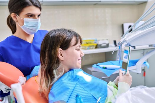 Teenage female looking at healthy teeth in mirror, in dental office, visit to dentist orthodontist. Treatment and care of teeth, teenagers, orthodontics, dentistry concept