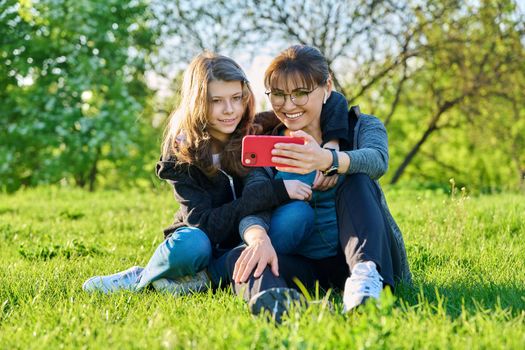 Mom and daughter 11, 12 years old having fun looking together at smartphone screen, outdoor, sitting embracing on grass. Family, leisure, lifestyle, relationship love, mother's day, motherhood concept