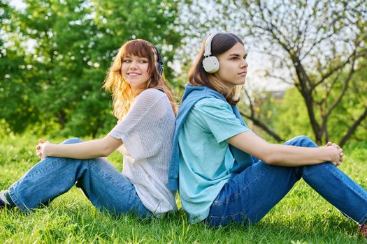 Two friends guy girl listen to music podcast, in wireless headphones, sitting on lawn on grass. Enjoying friendship, music, nature. Youth, lifestyle, young people concept