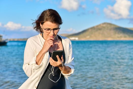 Happy 40s age woman in headphones with smartphone, sky sea nature. Beautiful female listening looking at smartphone screen walking relaxing on beach, copy space. Tourism, travel, vacation, technology