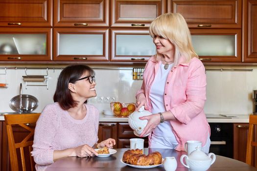 Two smiling happy mature women girlfriends in the kitchen together, having fun drinking tea chatting. Friendship, leisure, communication, 40s 50s age people concept