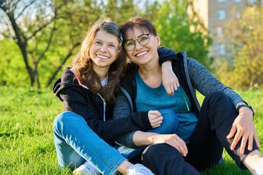 Happy mother and daughter hugging sitting on grass, looking at camera. Mom and girl resting together on lawn. Family, happiness, leisure, lifestyle, relationship love, mother's day, motherhood concept