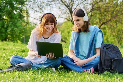 Two friends students, guy and girl in headphones with backpacks, looking at laptop, sitting on grass. High school, college, communication, friendship, education, youth concept