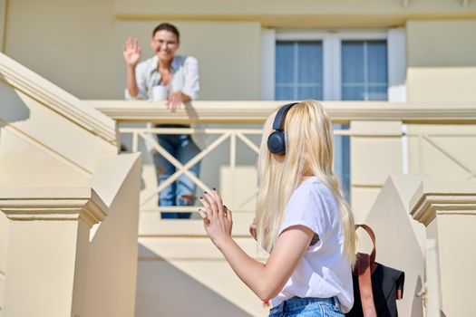 Middle-aged mother seeing off her teenage daughter on the porch of the house. Girl student in headphones with a backpack walking up the stairs. Family, lifestyle, parent and teenager concept