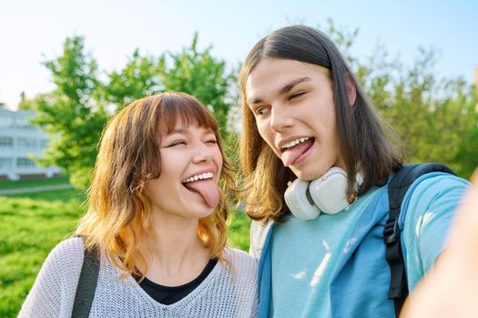 Young teenage friends guy and girl having fun, grimacing, making faces, laughing, taking selfie photo. Youth couple enjoy vacation, weekend, holiday, park sky background. Fun, joy, happiness concept