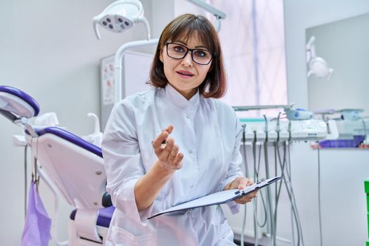 Talking consulting female doctor dentist looking at webcam, making video call chat conference, notes on clipboard, in dental office. Online technology, telemedicine, help medicine, health care concept