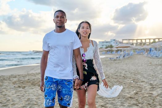 Young happy couple walking on beach holding hands. Multicultural couple in love together on sea shore. Honeymoon, dating, happiness, relationship, vacation, love, romance concept