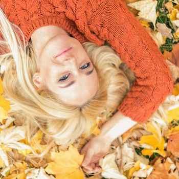 Beautiful young caucasian woman lying down on yellow leaves looking at camera in Autumn Park