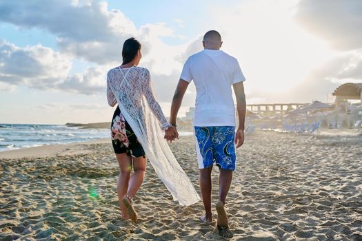 Young happy couple walking on the beach holding hands, back view. Honeymoon, dating, happiness, relationship, vacation, love, romance concept