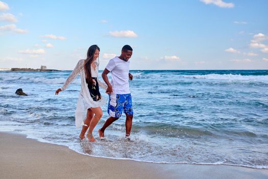 Young happy couple walking on beach holding hands, copy space. Multicultural couple in love together on sea shore. Honeymoon, dating, happiness, relationship, vacation, love, romance concept