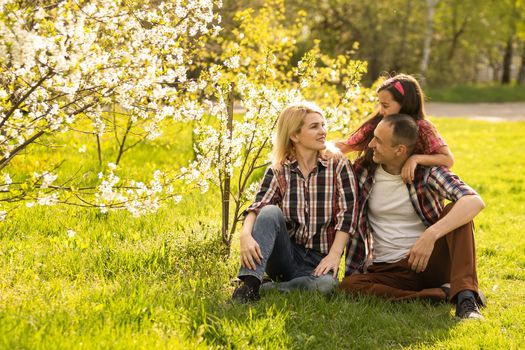 Outdoor portrait of happy young family playing in spring park under blooming tree, lovely couple with little child having fun in sunny garden.