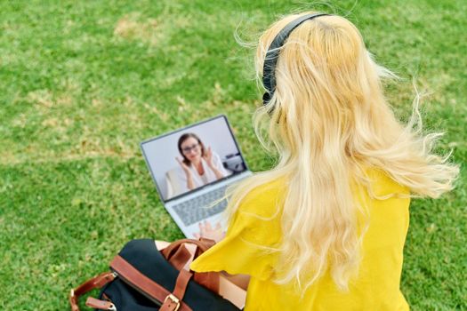 Remote online lecture on laptop screen, web chat with teacher individual consultation of teenage. College high school student girl sitting on grass in park, talking listening tutor on video conference