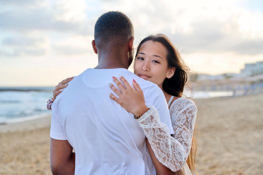 Young loving couple hugging on the beach. Face of beautiful Asian woman, man back. Love, relationship, honeymoon, vacation together, happiness, romantic, travel, family concept