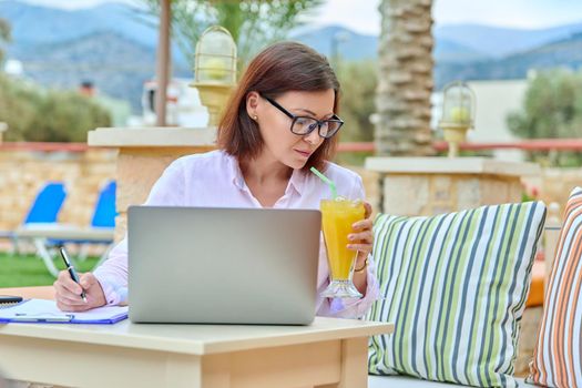 Beautiful business woman in her 40s working on a laptop remotely, sitting at an outdoor table in a resort hotel. Freelance work, business, middle aged people concept