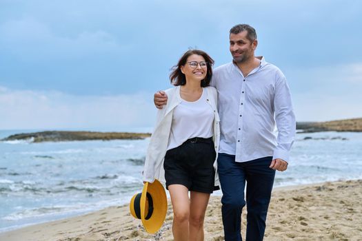 Happy mature couple walking on the beach. Smiling talking middle-aged man and woman embracing together on the seashore. Age, relationship, love, leisure, vacation, people concept