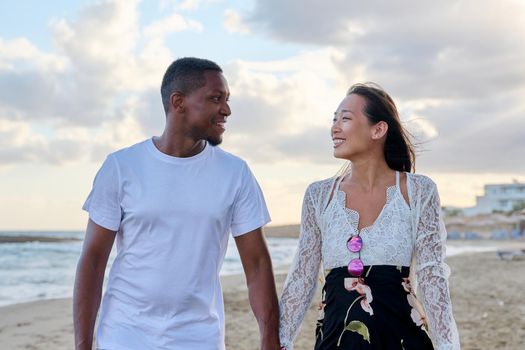 Young happy couple walking on beach holding hands. Multicultural couple in love together on sea shore. Honeymoon, dating, happiness, relationship, vacation, love, romance concept