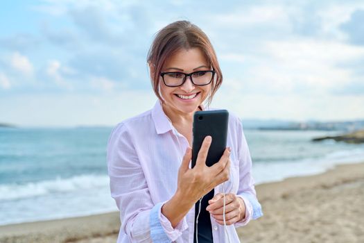 Happy middle aged woman on sea vacation enjoying in headphones with smartphone in her hands. Vacation, lifestyle, technology, leisure, people 40s concept