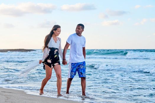 Young happy couple walking on beach holding hands, copy space. Multicultural couple in love together on sea shore. Honeymoon, dating, happiness, relationship, vacation, love, romance concept