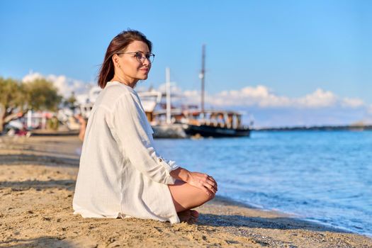 Mature woman sitting in lotus position meditating on beach. Female enjoys sea nature, setting sun, summer vacations. Lifestyle, tourism, travel, beauty, health, relaxation, mature people concept