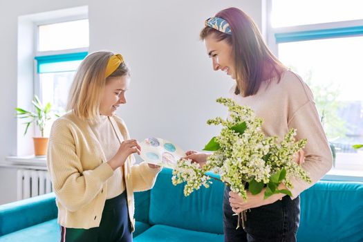 Happy mother and daughter child congratulating with bouquet of flowers and hand-drawn card, at home in living room. Mother's day, family, relationship, emotions, love, togetherness concept