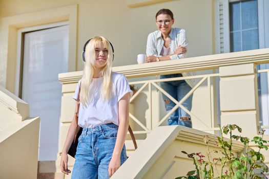 Middle-aged mother seeing off her teenage daughter on the porch of the house. Girl student in headphones with a backpack walking up the stairs. Family, lifestyle, parent and teenager concept