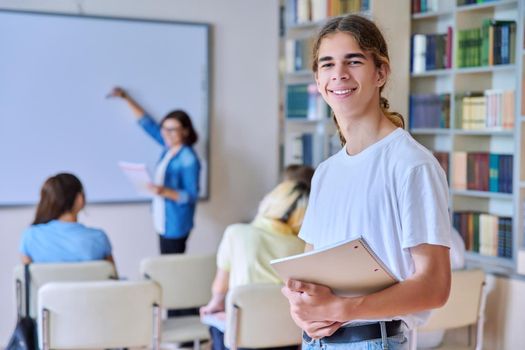 Teenage guy in classroom looking at camera, group of high school students with teacher study in library. Education, learning, knowledge, adolescence concept