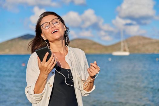 Happy middle aged woman in headphones listening to music using smartphone on beach. Mature female with closed eyes enjoying music, sunset, sea holidays. Vacation, feelings, nature, people 40s concept