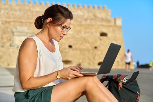 Mature woman tourist using laptop, old european historic fortress background. Technology, vacations, tourism, leisure, active lifestyle, middle aged people concept