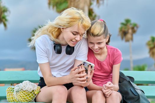 Friendly family, two girls sisters, teenage and preteen look at smartphone. Sitting in park with tropical trees, summer vacation together