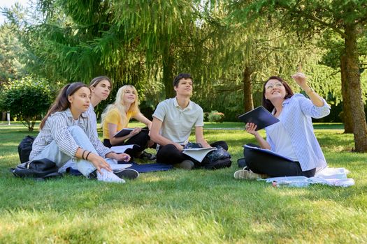 Outdoor, group of students with female teacher. Teenagers and mentor teacher talking sitting on grass in college park. Back to school, back to college, high school, education, teenagers concept