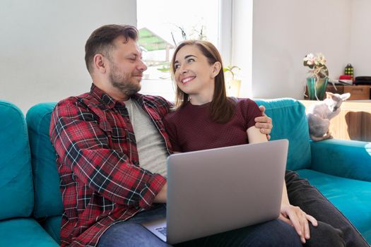 Happy smiling middle aged family couple looking at laptop screen together while sitting on sofa at home with pet cat. Relationships, lifestyle, family, leisure, technology, rest, people 40s concept