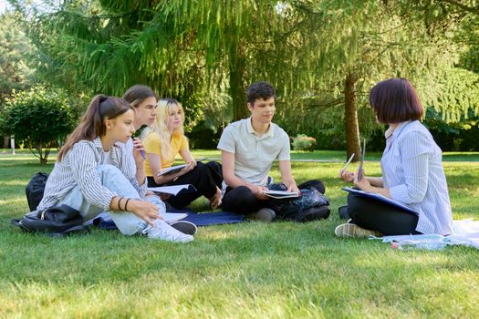 Outdoor, group of students with female teacher. Teenagers and mentor teacher talking sitting on grass in college park. Back to school, back to college, high school, education, teenagers concept