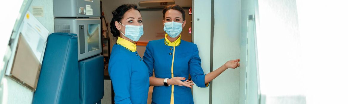 Female flight attendants wearing protective face masks while standing near airplane door and welcoming passengers